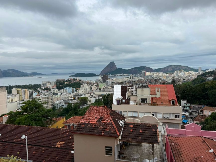 Una amplia vista de la ciudad que contempla una bahía costera con edificios densos, tejados de tejas rojas y colinas verdes bajo un cielo nublado. En la distancia, una montaña dramática se eleva sobre el agua, dando a la escena una sensación melancólica y expansiva.