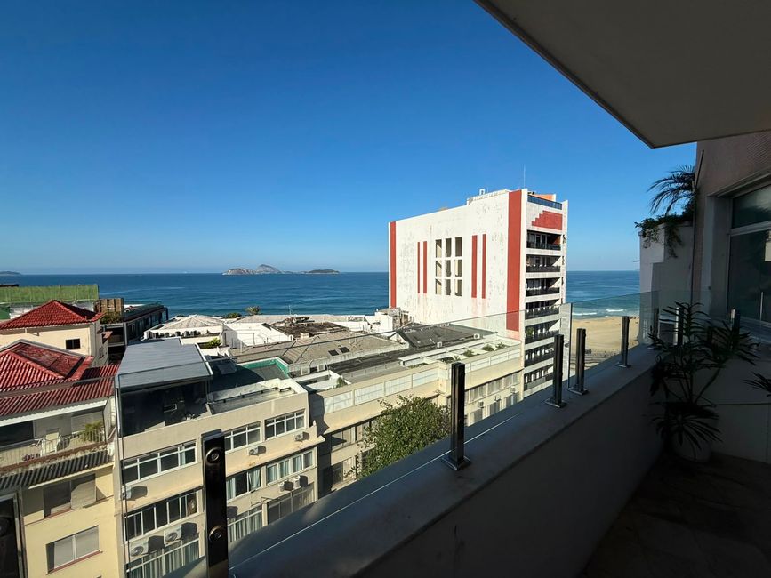 Una vista brillante del mar desde un balcón que da a edificios bajos, una playa de arena y el océano azul. La escena se siente tranquila y soleada, con un cielo despejado y islas distantes en el horizonte.