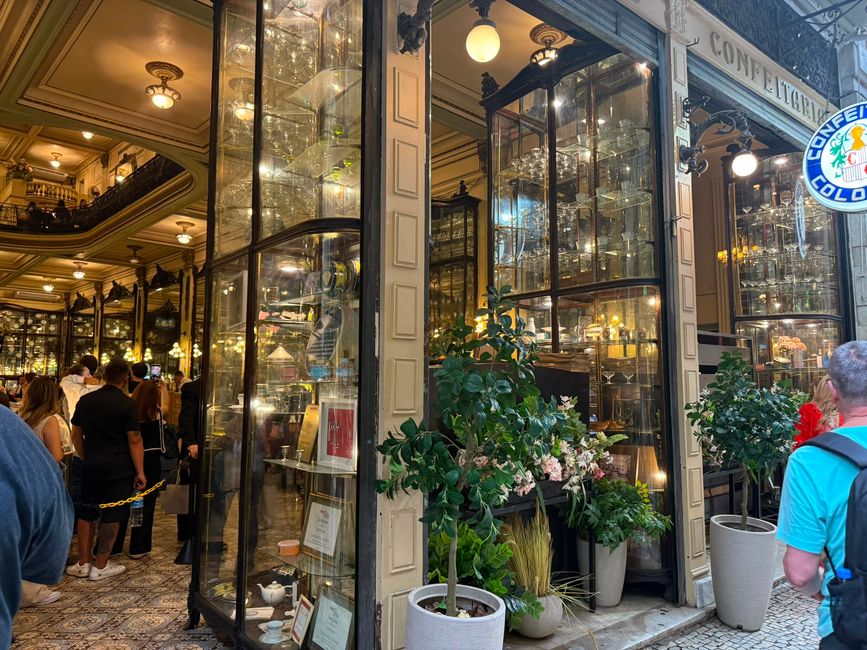 A busy, elegant café or confectionery with ornate glass display cabinets and warm lighting, crowded with people inside and outside. The scene feels lively and upscale, with potted plants and a classic old-world interior.