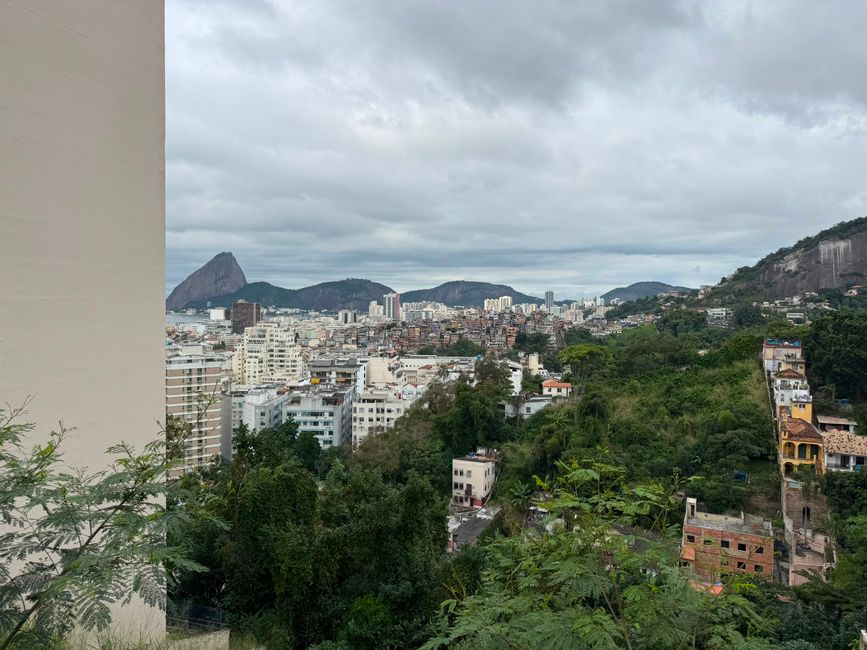 Una amplia vista de la ciudad con edificios densos, colinas verdes y un dramático pico rocoso a lo lejos bajo un cielo nublado. La escena se siente tranquila y nublada, con vegetación exuberante en primer plano.