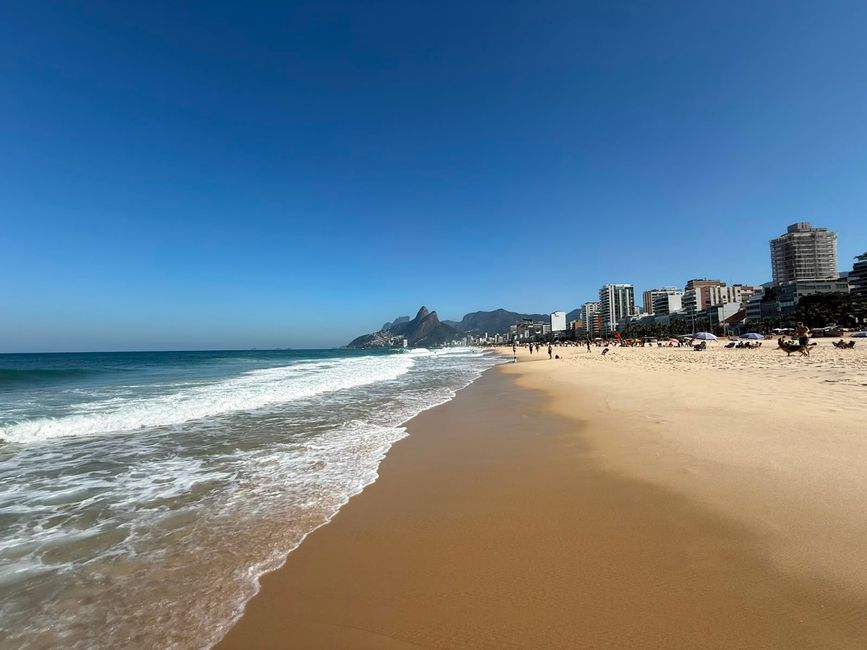 Una amplia playa de arena con suaves olas que rompen bajo un cielo azul claro, con un horizonte donde se pueden ver la línea del horizonte de la ciudad y montañas a lo lejos. La escena se siente brillante, soleada y relajada, con personas dispersas a lo largo de la costa.