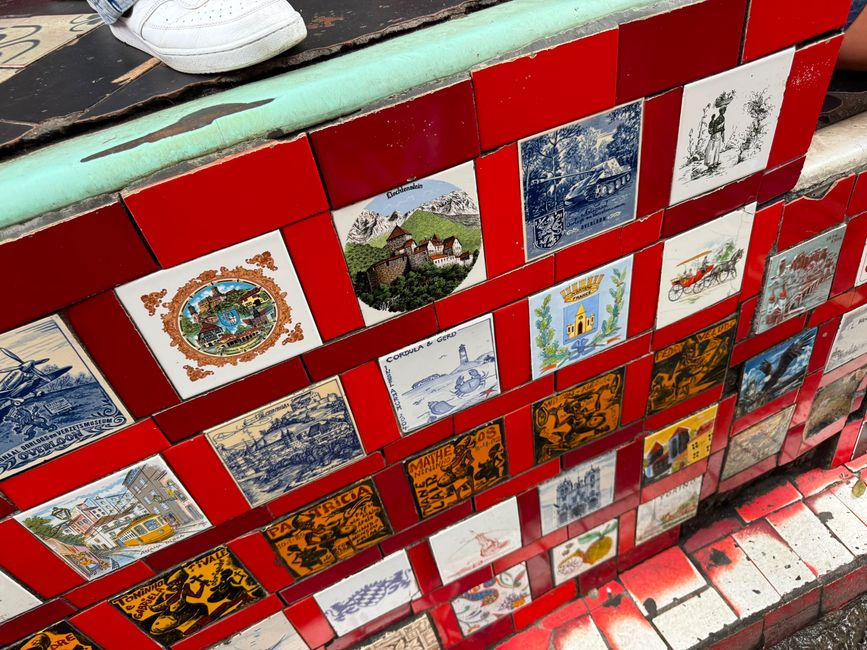 Una colorida pared de escalera cubierta de azulejos de viaje y souvenirs de estilo vintage, con escalones rojos brillantes y un borde desgastado en color turquesa. Una zapatilla blanca en la parte superior sugiere que alguien está subiendo por las escaleras decorativas.