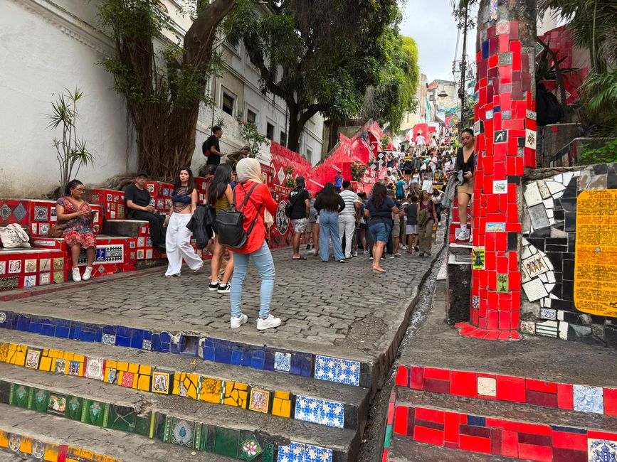 Una escalera colorida y vibrante cubierta de azulejos rojos y estampados está llena de visitantes explorando y tomando fotos. La escena se siente ocupada y vibrante, con árboles y edificios urbanos enmarcando la empinada calle.