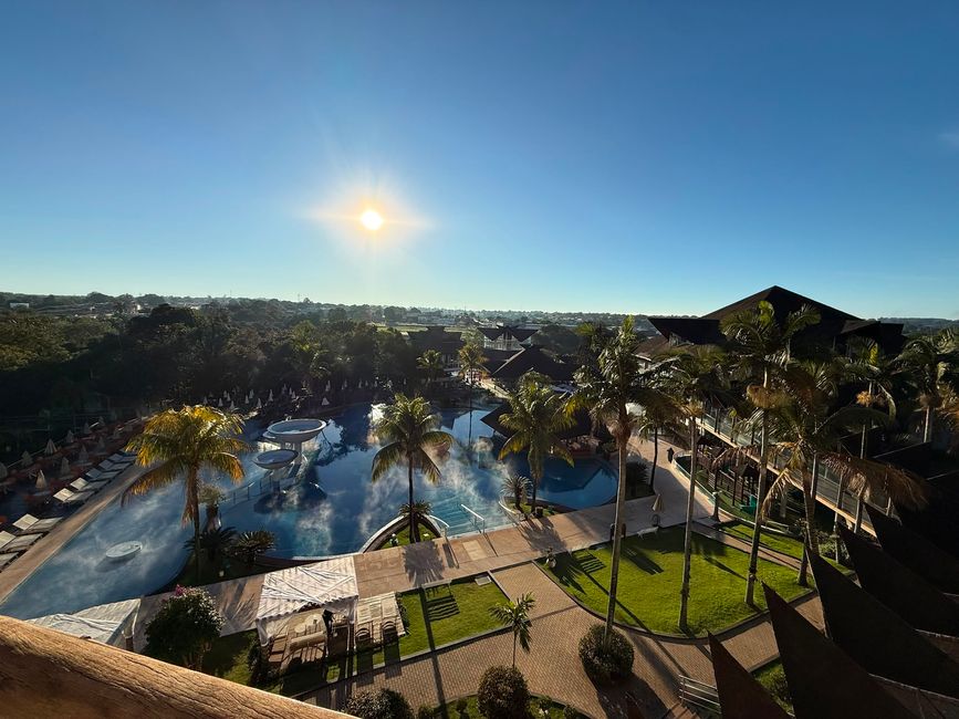 Una escena de resort soleada con una gran piscina, palmeras y áreas de descanso bajo un cielo azul claro. La cálida luz de la mañana le da al lugar un ambiente relajado y tropical.