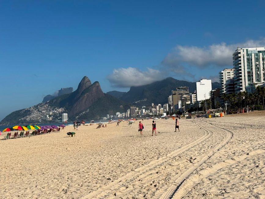 Una escena de playa soleada con amplia arena, sombrillas coloridas y personas caminando a lo largo de la costa. En el fondo, altos edificios de la ciudad y dramáticas montañas verdes se elevan bajo un cielo azul claro, creando un ambiente tropical relajado.