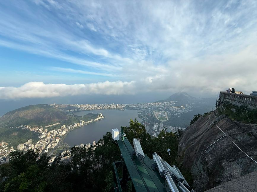 Una amplia vista desde la cima de una montaña sobre una ciudad costera, con una bahía, barrios densos y nubes dramáticas que se extienden por el cielo. Unas pocas personas están de pie en un mirador rocoso, dando a la escena una sensación de calma y un viaje escénico.