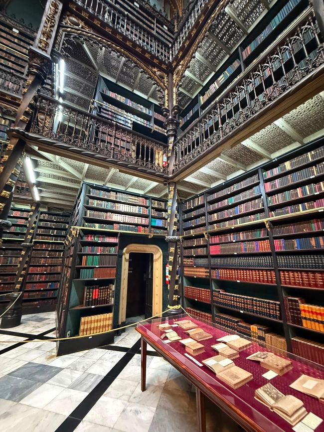 An ornate multi-level library filled with rows of old books and decorative iron balconies. The quiet interior feels grand and historic, with display tables of rare books in the foreground.