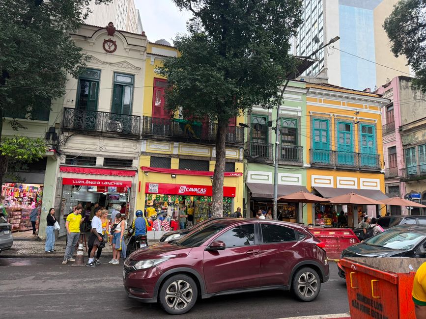A lively city street with colorful old buildings, balconies, and shops at ground level. People gather on the sidewalk while cars pass by, giving the scene a busy, vibrant atmosphere.