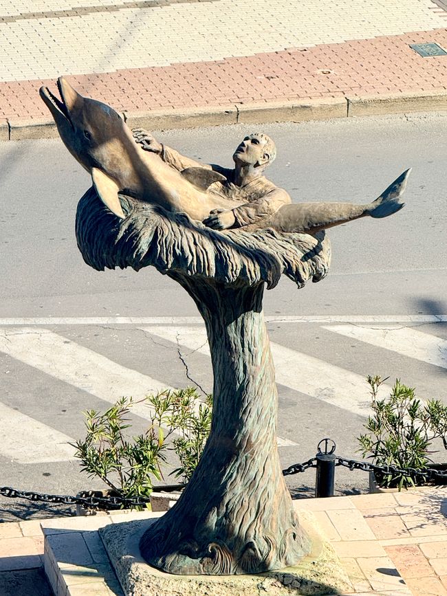 Ein Delfin rettet einen Seemann aus den Fluten – eine Statue, die in den Hafen passt.