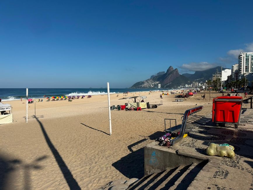 Una escena de playa soleada con amplia arena dorada, olas que rompen y coloridas sombrillas a lo largo de la costa. A lo lejos, una montaña dramática se eleva junto al horizonte de la ciudad, dando a la vista una sensación costera relajada pero vibrante.
