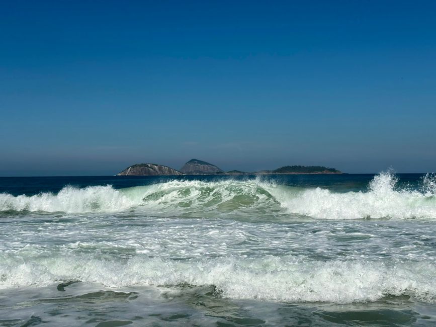 Olas poderosas rompen en una playa de arena bajo un cielo azul claro, con pequeñas islas rocosas visibles en el horizonte. La escena se siente brillante, abierta y refrescante.