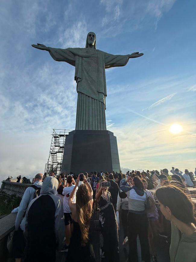 Una gran multitud se reúne a los pies de la estatua de Cristo Redentor, con muchas personas tomando fotos bajo un cielo brillante al atardecer. La escena se siente bulliciosa e inspiradora, con nubes y luz cálida enmarcando el monumento.