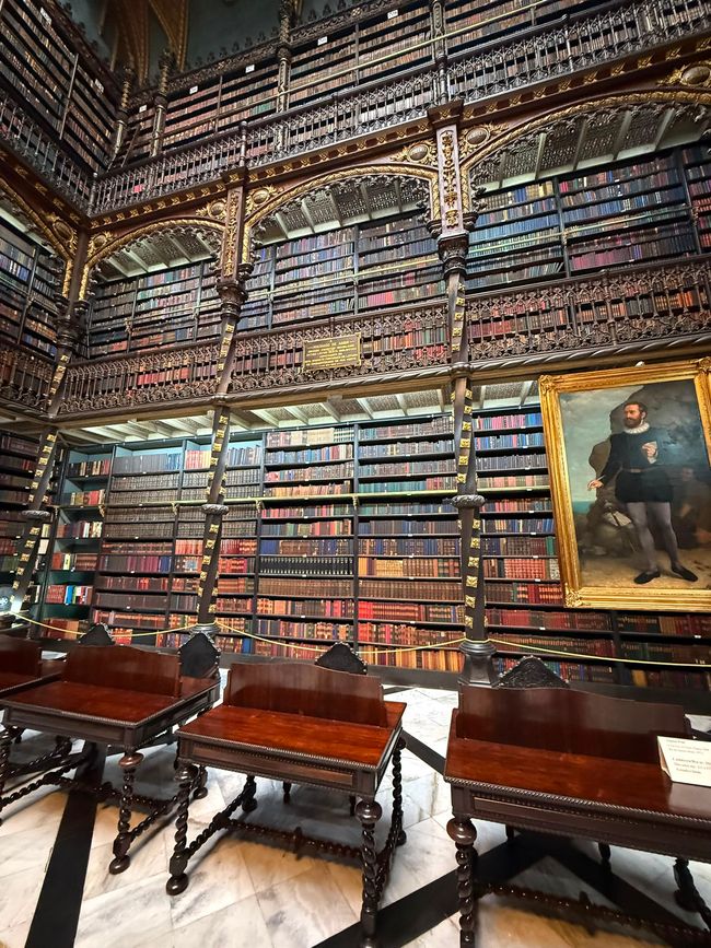 An ornate historic library with towering bookshelves, carved balconies, and rows of wooden reading tables. The grand, dimly lit interior feels quiet and impressive, with a large portrait adding to the old-world atmosphere.