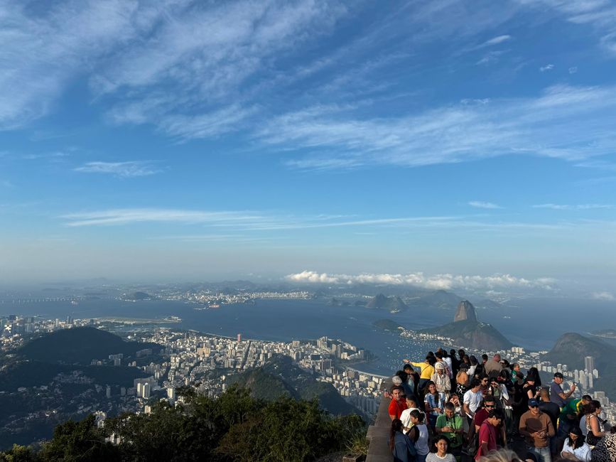 Un mirador concurrido contempla una extensa ciudad costera, una bahía y picos graníticos icónicos bajo un cielo azul brillante. La escena se siente animada y escénica, con visitantes reunidos para disfrutar de la vista panorámica.