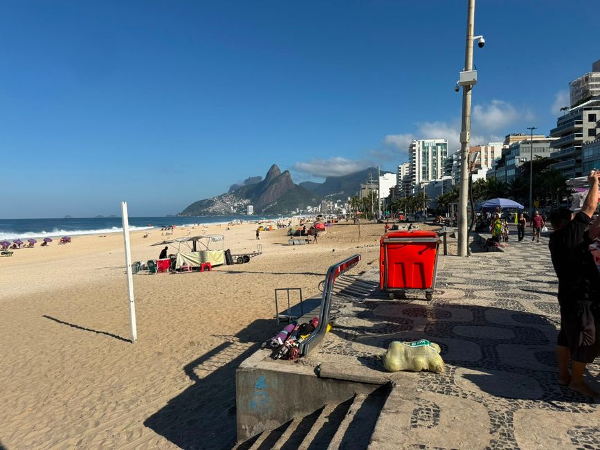 Un paseo marítimo soleado corre paralelo a una amplia costa de arena, con olas, bañistas dispersos y edificios de la ciudad que se extienden a lo lejos. Montañas se elevan en el fondo bajo un cielo azul claro, dando a la escena una agradable sensación costera de relax.