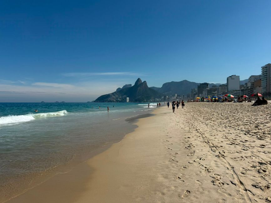 Una escena de playa soleada con suaves olas, personas caminando y nadando, y un horizonte de ciudad respaldado por montañas dramáticas a lo lejos. La amplia costa de arena y el cielo azul claro le dan un aire relajado y animado de vacaciones.