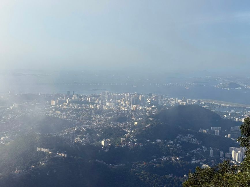 Una vista panorámica brumosa de una ciudad costera con edificios densos, colinas verdes y un largo puente que se extiende a través del agua. La escena se siente tranquila y expansiva, vista desde un alto mirador.