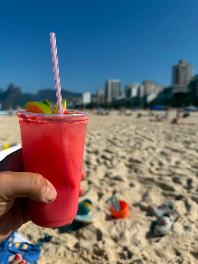 Una mano sostiene una bebida helada de color rosa brillante con una pajita y un adorno de lima en una playa soleada. En el fondo, la costa de arena, artículos de playa dispersos y un horizonte de ciudad borroso crean un ambiente vacacional relajado.