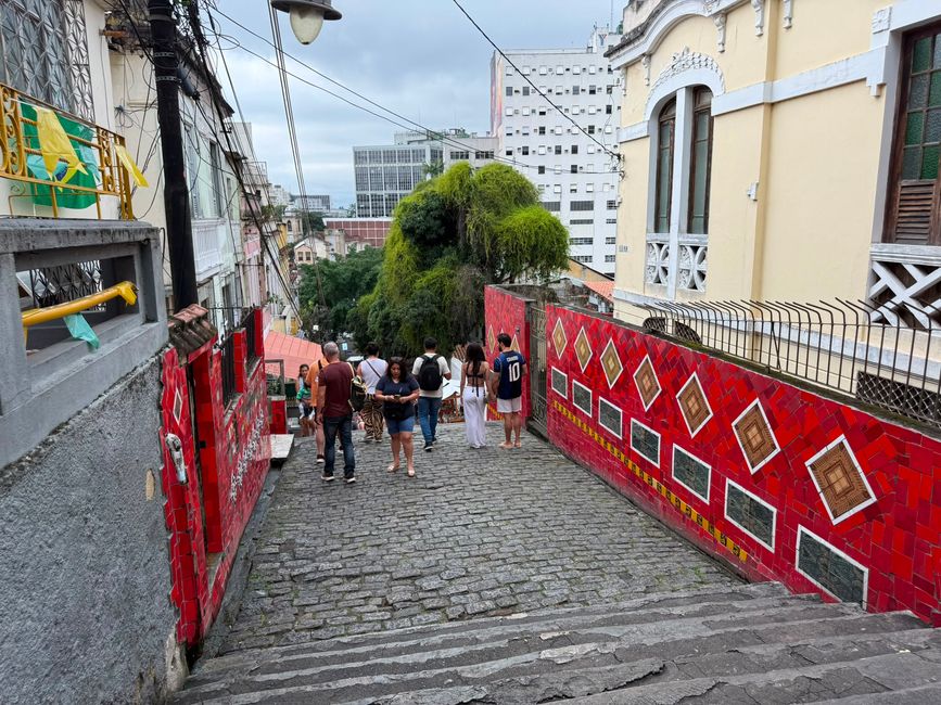 Las personas caminan por una empinada calle adoquinada flanqueada por paredes de azulejos rojos brillantes y viejos edificios, con el horizonte de la ciudad y un cielo nuboso al fondo. La escena se siente animada y colorida, con una mezcla de encanto histórico y energía urbana.