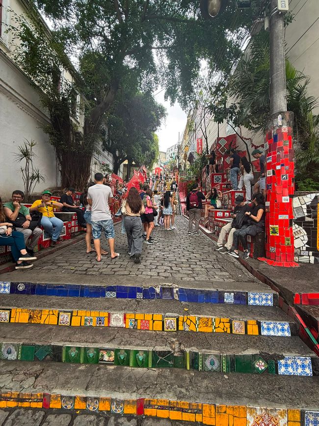 Una escalera de calle vibrante y colorida cubierta de azulejos de mosaico, con muchas personas sentadas, caminando y tomando fotos. Altos árboles se arquean sobre la escena, dándole una vibra urbana de viaje ocupada pero relajada.