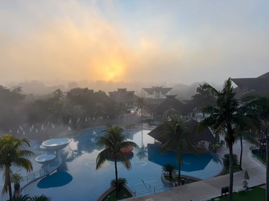 Un amanecer brumoso brilla sobre un complejo turístico tropical con palmeras, piscinas y edificios de techos oscuros. La escena se siente tranquila y soñadora, con una suave luz reflejándose en el agua.